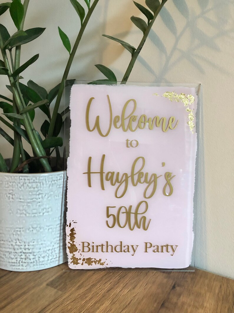 May include: A pink and gold welcome sign with the text "Welcome to Hayley's 50th Birthday Party".