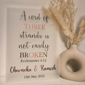 May include: A frosted glass sign with the text "A cord of THREE strands is not easily BROKEN Ecclesiastes 4:12 Cherneka & Ramish 15th May 2022" in rose gold lettering. The sign is on a white background with a vase of dried flowers to the right.