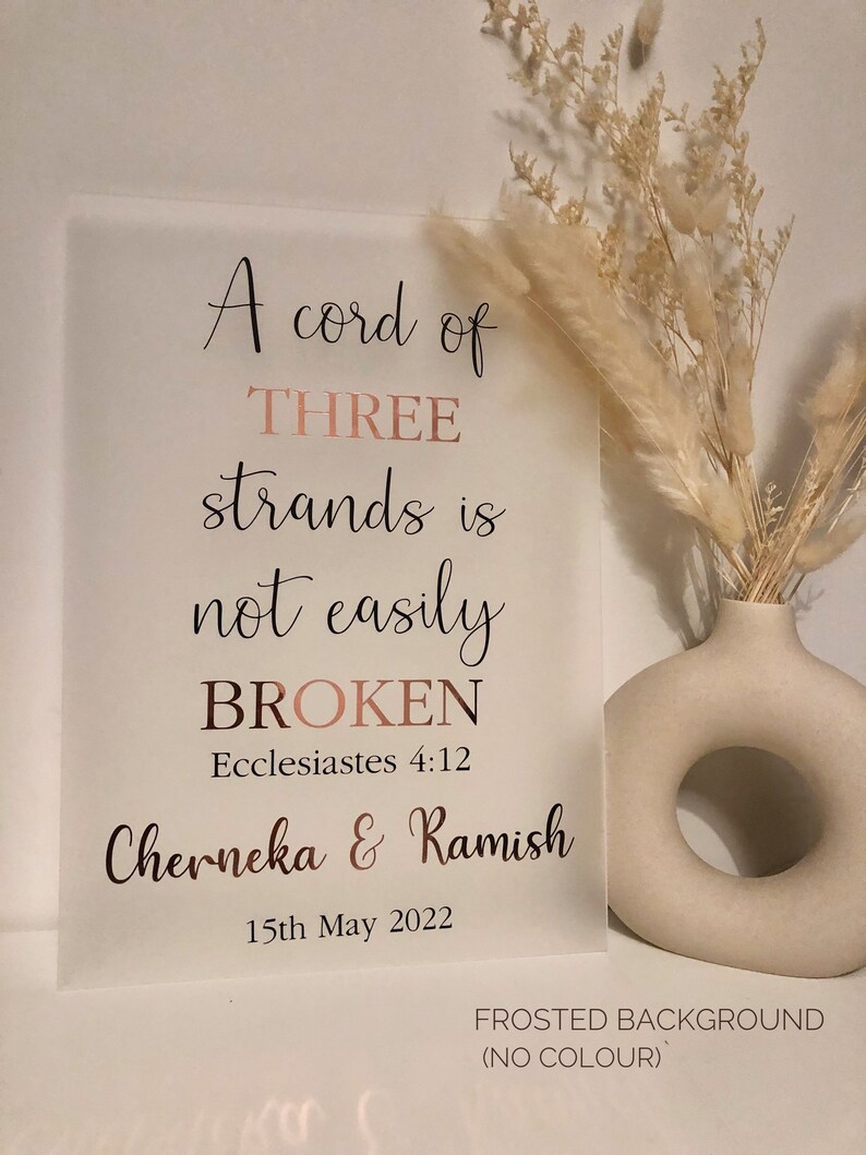 May include: A frosted glass sign with the text "A cord of THREE strands is not easily BROKEN Ecclesiastes 4:12 Cherneka & Ramish 15th May 2022" in rose gold lettering. The sign is on a white background with a vase of dried flowers to the right.