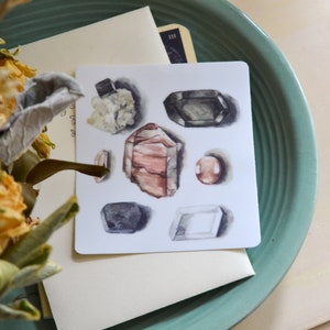 Puede incluir: Una tarjeta blanca con ilustraciones de seis gemas diferentes. Las gemas están dibujadas en acuarela e incluyen una gema rosa, una gris, una blanca, una negra y dos gemas verdes.