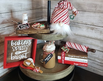 Può includere: Un vassoio a tre livelli decorato per Natale con un cartello rosso che dice "Mrs. Claus Bakery" e un gnomo che indossa un cappello a righe rosse e bianche. Il vassoio include anche un barattolo di caffè, un mattarello e un cucchiaio di legno.