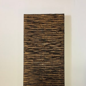 Op de afbeelding: Een rechthoekig houten wandkunstwerk met een getextureerd oppervlak. Het kunstwerk heeft een patroon van horizontale lijnen in bruin- en zwarttinten, waardoor een natuurlijke, rustieke esthetiek ontstaat. Het stuk wordt gepresenteerd tegen een witte achtergrond.