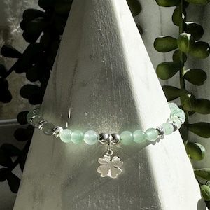 May include: A green jade stone bracelet with a silver four-leaf clover charm. The bracelet is displayed on a white marble cone.