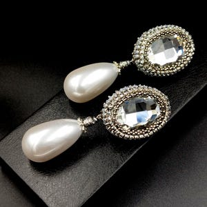 May include: A pair of drop earrings. Each earring has a large, pearlescent teardrop-shaped bead suspended below an oval, clear crystal surrounded by a silver-toned beaded frame. The earrings are set against a black background.