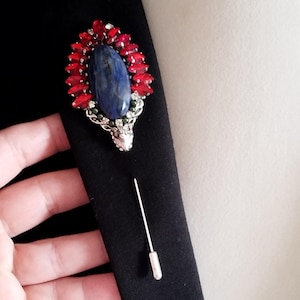 May include: A silver brooch with a blue stone surrounded by red stones. The brooch is attached to a silver pin.