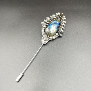 May include: A silver pin with a teardrop-shaped iridescent blue gemstone surrounded by silver and black rhinestones.