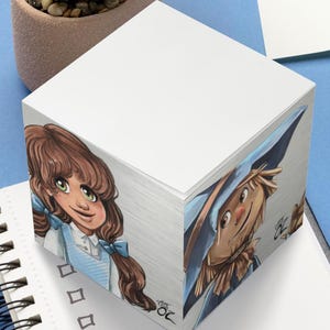 May include: A white cube notepad featuring illustrations of characters from The Wizard of Oz. A small succulent plant in a brown pot and a spiral-bound notebook are on a blue surface.