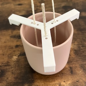 May include: A white plastic candle wick centering tool with three slots for wicks. The tool is placed inside a light pink ceramic candle jar.