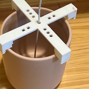 Puede incluir: Portavelas de plástico blanco con cuatro brazos, diseñado para mantener una mecha de vela en su lugar. El soporte se coloca dentro de un recipiente de cerámica rosa claro.