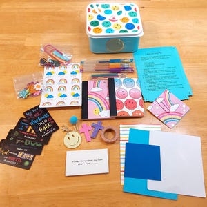 May include: A collection of stationery and craft supplies, including notebooks, pens, stickers, and decorative tape. The items feature smiley faces, rainbows, and inspirational quotes. A blue box with a smiley face lid is also present.