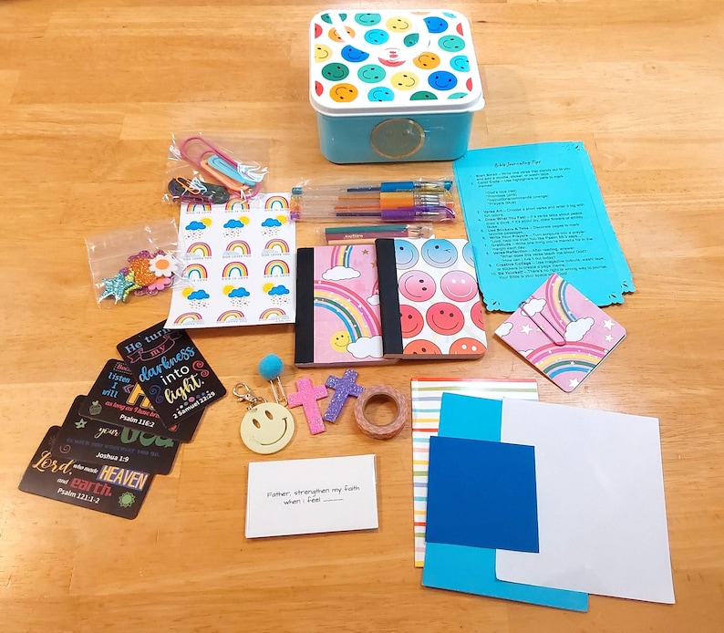 May include: A collection of stationery and craft supplies, including notebooks, pens, stickers, and decorative tape. The items feature smiley faces, rainbows, and inspirational quotes. A blue box with a smiley face lid is also present.