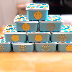 May include: A stack of light blue rectangular containers with white lids. The lids are decorated with colorful polka dots. Each container has a gold smiley face or circle on the front.