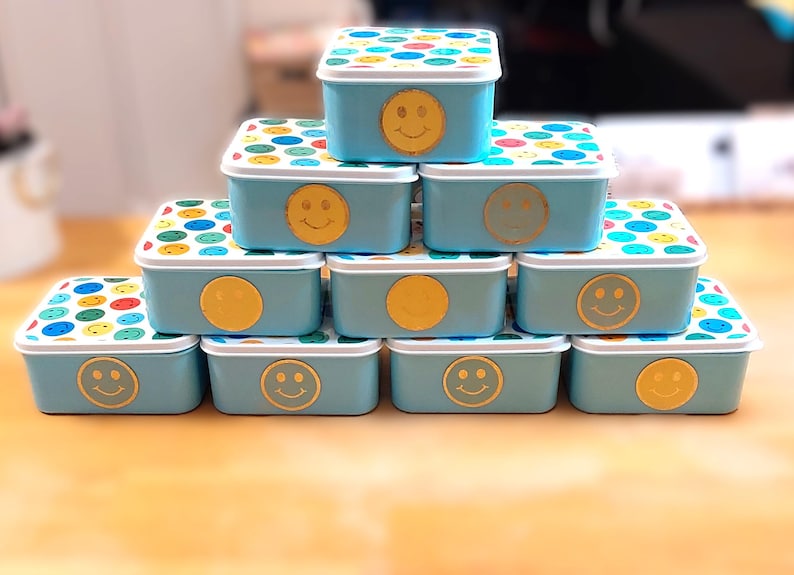 May include: A stack of light blue rectangular containers with white lids. The lids are decorated with colorful polka dots. Each container has a gold smiley face or circle on the front.