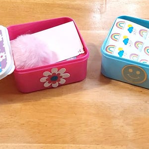 May include: Two open rectangular boxes with lids. One is pink with a daisy and contains a pink puff and a white card. The other is light blue with a smiley face and contains a rainbow-patterned card. The lids have colorful patterns.