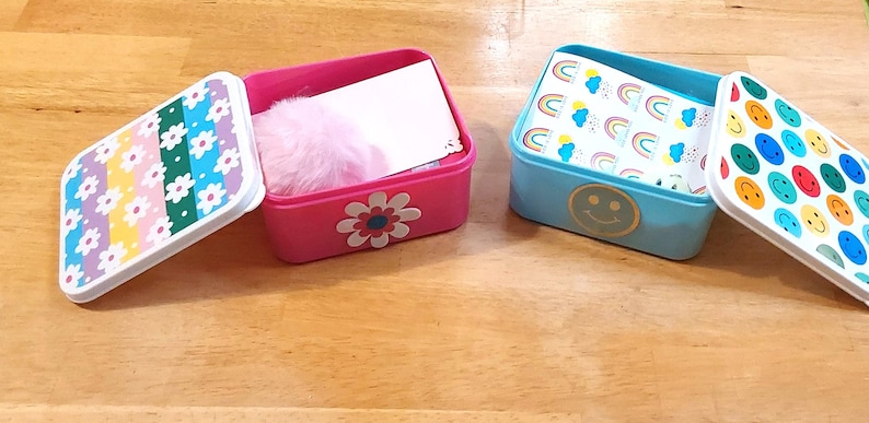May include: Two open rectangular boxes with lids. One is pink with a daisy and contains a pink puff and a white card. The other is light blue with a smiley face and contains a rainbow-patterned card. The lids have colorful patterns.