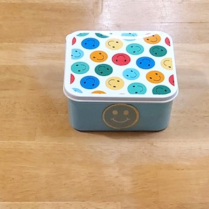 May include: A small, square, light blue metal box with a white rim and a lid covered in colorful smiley faces. A gold smiley face is on the front of the box. The box is sitting on a wooden surface.