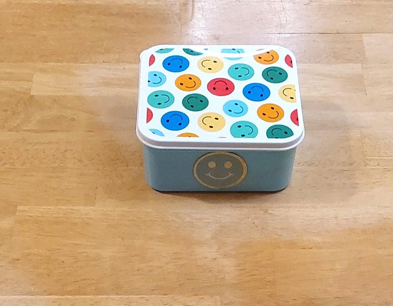 May include: A small, square, light blue metal box with a white rim and a lid covered in colorful smiley faces. A gold smiley face is on the front of the box. The box is sitting on a wooden surface.