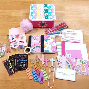 May include: A collection of journaling supplies, including a pink floral box, notebooks, pens, stickers, and paper clips. The notebooks feature rainbow and star designs. The box is pink with a floral design.