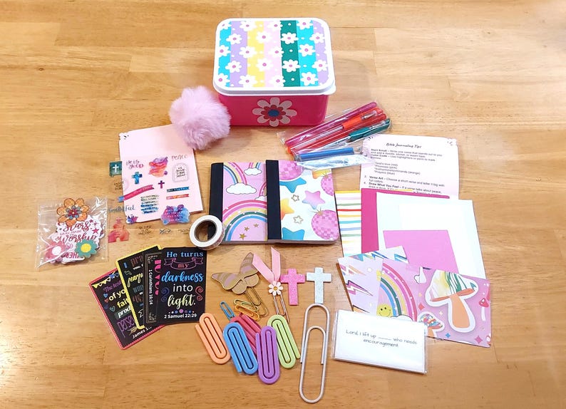 May include: A collection of journaling supplies, including a pink floral box, notebooks, pens, stickers, and paper clips. The notebooks feature rainbow and star designs. The box is pink with a floral design.