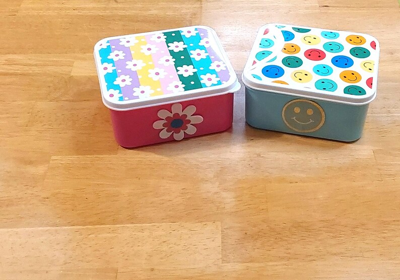 May include: Two rectangular lunch boxes. One is pink with a floral design and a large flower on the front. The other is light blue with a smiley face and a lid covered in colorful smiley faces. Both have white rims.