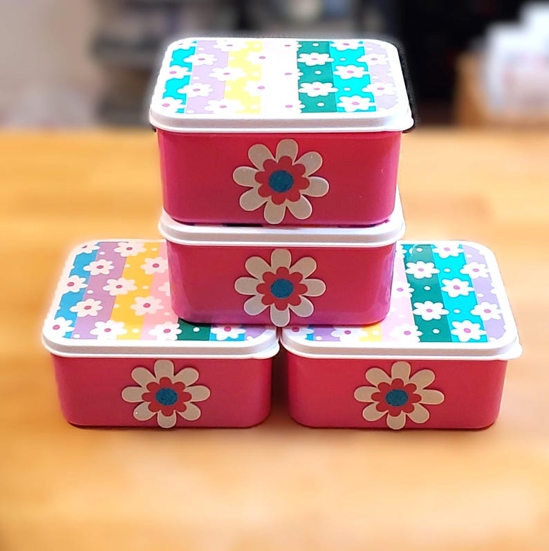 May include: Four pink rectangular storage containers stacked. Each container has a white lid with a floral pattern in pastel colors. A large flower with a blue center decorates the front of each container. The containers are on a wooden surface.