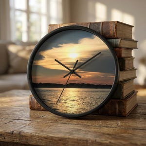 Puede incluir: Un reloj de pared redondo negro con una imagen de puesta de sol. Las manecillas del reloj son negras. El reloj está sobre una superficie de madera junto a una pila de libros antiguos con cubiertas marrones.