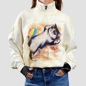 May include: Cream-colored sweatshirt featuring a graphic of a cat wearing sunglasses, lounging on a beach. A colorful beach ball and a small red umbrella are also depicted. The cat is black and white. The sweatshirt has a high collar.