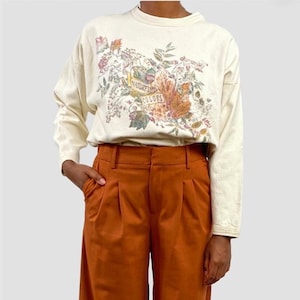 May include: Cream-colored long-sleeve sweatshirt with a floral graphic and the text "Nature's Colors." The sweatshirt is paired with rust-colored pleated pants. The outfit is casual and features a nature-inspired design.