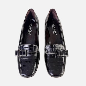 May include: A pair of black leather loafers with a crocodile texture. The shoes have a square toe, a buckle detail, and white stitching. The text "MILA PAOLI MADE IN ITALY" is visible on the inner sole.