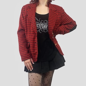 May include: A red and black houndstooth blazer with gold buttons. The blazer is worn over a black tank top with the text "EY-DAV" and a black pleated skirt. The outfit is completed with patterned tights.