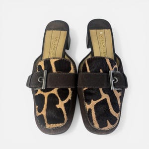 May include: A pair of slip-on mules featuring a brown suede finish and a textured animal print panel on the vamp. Each shoe includes a decorative brown suede strap across the front with a silver-toned metal buckle. The insole is labeled Villager.