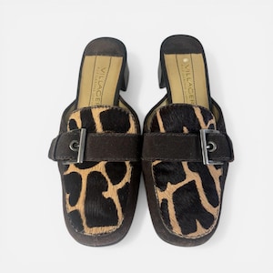 May include: A pair of brown leather slip-on shoes with a giraffe print design on the front. The shoes have a dark brown strap with a silver buckle. The brand name "VILLAGER" is printed on the insole.
