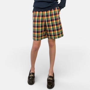 May include: A person wearing a navy blue sweater and plaid shorts in shades of yellow, green, red, and brown. The person is wearing black loafers with gold chain detailing.
