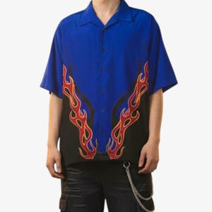 May include: A short-sleeved button-down shirt with a blue upper section and a black lower section. The shirt features a flame design in red and yellow, creating a bold visual contrast. The shirt has a classic collar and a relaxed fit.