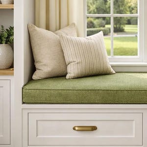 May include: A white window seat with two storage drawers featuring brass handles, topped with a green cushion and four throw pillows in solid and striped beige fabric, positioned beneath a window with cream curtains and a side table with a lamp.