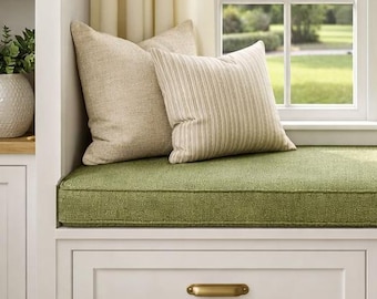 May include: A white window seat with two storage drawers featuring brass handles, topped with a green cushion and four throw pillows in solid and striped beige fabric, positioned beneath a window with cream curtains and a side table with a lamp.