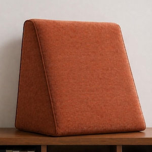 May include: A terracotta-coloured, triangular cushion with a textured finish. It sits on a wooden shelf, with books and a decorative sphere below. A window with greenery is visible in the background.