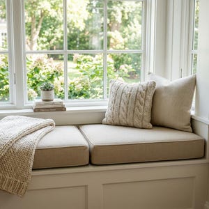 May include: A window seat featuring a beige upholstered cushion, a chunky cable-knit throw blanket, and two decorative pillows. One pillow has a textured cable-knit pattern, while the other is plain beige. A small potted succulent sits on books.