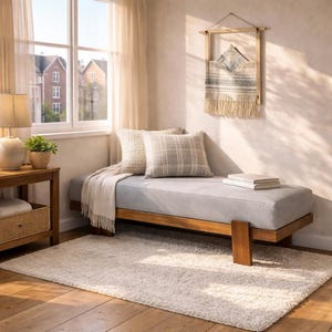 May include: A daybed with a light gray cushion and a wooden frame, featuring a cream-colored throw blanket, a textured cream pillow, and a gray plaid pillow. A wall hanging with fringe and a stack of books sit nearby on the daybed surface.