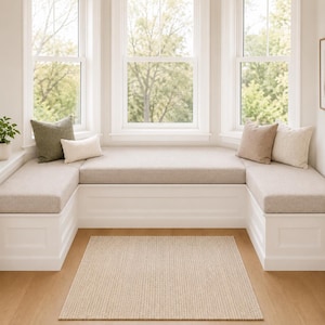 May include: A white U-shaped built-in window seat with beige upholstered cushions, featuring four decorative throw pillows in olive green, cream, and tan colors, positioned in front of three large windows with a textured beige area rug below.