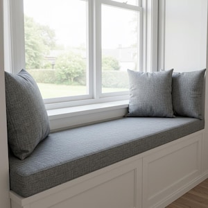 May include: A window seat with a gray cushion and two matching pillows. The seat is built into a white frame with paneled detailing. The window behind the seat offers a view of a green yard and trees.