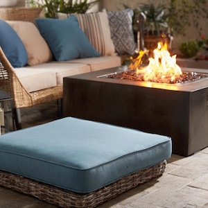 May include: Outdoor patio scene featuring a wicker sofa with blue and beige cushions, a square fire pit with flames, and a woven ottoman with a blue cushion. The scene evokes a relaxing, comfortable atmosphere.