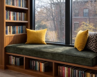 May include: A built-in wooden window seat featuring a dark green velvet cushion, two solid mustard yellow throw pillows, and one gray patterned pillow. The unit includes integrated shelving filled with books above and below the seating area.
