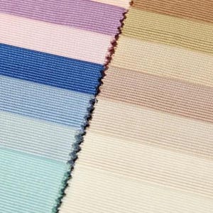 Puede incluir: Una colección de muestras de tela dispuestas en dos columnas verticales. La columna izquierda presenta tonos lavanda, rosa, azul real, azul claro y verde azulado. La columna derecha muestra tonos neutros que incluyen beige y crema, todos con textura acanalada.