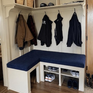 Könnte beinhalten: Eine maßgefertigte Eingangsbank mit marineblauem Kissen und Schuhaufbewahrung. Die weiße Bank hat ein eingebautes Regal für Schuhe und Haken zum Aufhängen von Mänteln und Jacken. Körbe und Hüte befinden sich auf dem Regal darüber.