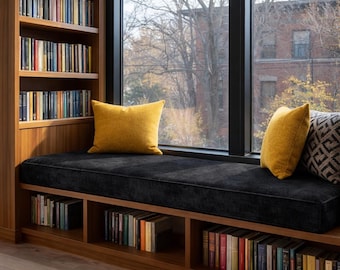 May include: A built-in wooden window seat featuring a black upholstered cushion, two solid mustard yellow throw pillows, and one patterned gray and black pillow. The unit includes integrated shelving filled with books above and below the seat.