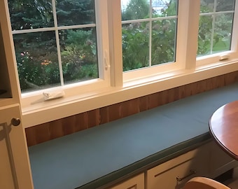 May include: A long, rectangular window seat cushion in a solid muted teal color, positioned atop a white built-in storage bench with drawers. The bench is situated directly beneath a three-pane window with white frames and a wood-paneled wall.