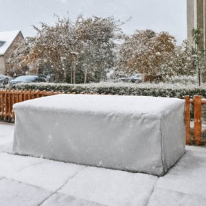 May include: A rectangular outdoor furniture cover in a light grey colour, draped over a large piece of furniture. The cover is made of a smooth, solid material and is completely blanketed in a layer of fresh white snow during a winter snowfall.