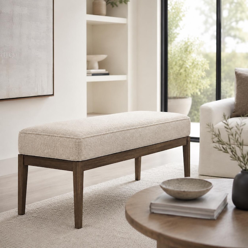 May include: A rectangular bench featuring a thick, beige upholstered seat cushion with a textured fabric finish. The bench is supported by a dark brown wooden frame with four straight, slightly tapered legs that provide a stable base.