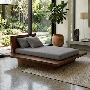 May include: A modern wooden daybed with a low profile. The bed features a dark wood frame, a gray cushion, and two gray pillows. The daybed is placed on a light brown rug in a well-lit room with a large window.
